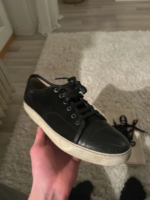 Svarta sneakers från Lanvin i skinn och mocka - Tjena! Säljer ett par svarta Lanvin sneakers med ovandel i mocka och tåparti i skinn. Skorna har rund tå, svarta snören och en vit platt sula med Lanvin-logga undertill. Klassisk och stilren modell som passar till det mesta. Har box och allt sådant, har växt ut skorna och vill bli av med dom därför det låga priset, har inget kvitto. Hör av er för frågor och funderingar!