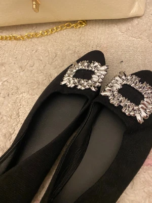 Svarta ballerinaskor med strassdetalj - Snygga svarta ballerinaskor med spetsig tå och stora glittriga strasspännen framtill. Skorna har en elegant och klassisk look, perfekta för att lyfta vilken outfit som helst. Materialet är textil med en mjuk känsla och insidan är svart.