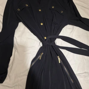 Svart klänning med gulddetaljer Michael Kors - Svart långärmad klänning från Michael Kors i storlek L. Klänningen har snygga guldfärgade knappar, två bröstfickor, dragkedjor vid höfterna och ett bälte i midjan med metallringar. Materialet är mjukt och följsamt, perfekt för dig som gillar stilrena detaljer.