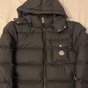 Svart Moncler pufferjacka med huva - Svart pufferjacka från Moncler i storlek S. Jackan har avtagbar huva, flera fickor med dragkedja och knapp, samt Moncler-logga på bröstet. Stilsäker look med quiltad design och hög krage. Perfekt för kalla dagar och riktigt snygg till streetwear. Den passar perfekt för de som har M också (jag har storlek M)