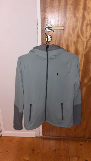 grön och grå vindjacka med huva från Peak Performance - Snygg grön och grå vindjacka från Peak Performance med huva och svarta dragkedjor. Jackan har två praktiska fickor framtill och diskret logga på bröstet. Perfekt för dig som gillar stilren och funktionell design. 