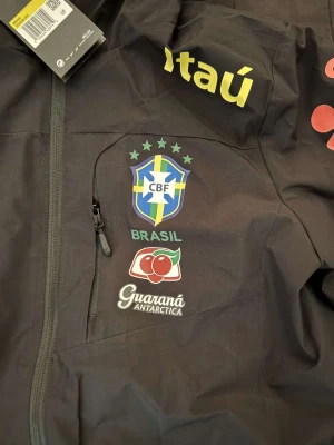 Svart Nike Brasil vindjacka med huva - Svart vindjacka från Nike med Brasil CBF-logga, sponsormärken som Itaú och Guaraná Antarctica. Jackan har huva, hel dragkedja och sportig look. Perfekt för dig som gillar fotboll.
