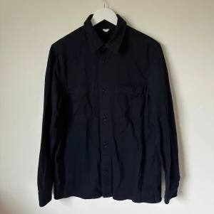 Mörkblå overshirt från Arket, strl 48 - Mörkblå overshirt från Arket i storlek 48. Skjortan har klassisk krage, två bröstfickor och knäppning framtill med svarta knappar. Materialet är mjuk bomull som känns skönt mot huden. Perfekt att bära som lager över t-shirt eller hoodie. 4Ö12