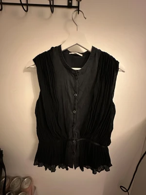 Svart plisserad blus från Vanessa Bruno - Svart blus från Vanessa Bruno med plisserade detaljer på sidorna och nedtill. Blusen är ärmlös, har rund hals och knäppning framtill med små svarta knappar. Materialet är tunt och lätt, vilket ger ett snyggt fall och en feminin siluett. En knapp är lite lös 