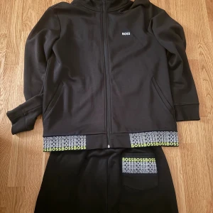 Svart hoodie från BOSS med dragkedja - Svart tracksuit från BOSS med hel dragkedja, huva och snörning med gröna detaljer. Långärmad och med fickor på sidorna. Nederkanten har BOSS-logga i vitt och grönt. Materialet är mjukt och känns som bomull eller polyester, perfekt för chill eller träning.