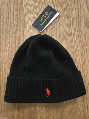 Svart kabelstickad mössa Polo Ralph Lauren - Svart kabelstickad mössa från Polo Ralph Lauren med bred ribbad kant och röd broderad logga framtill. Klassisk och stilren design som passar perfekt till kalla dagar. Materialet är mjuk och värmande ull.