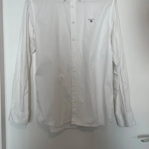Vit skjorta från GANT, 170 cm - Klassisk vit skjorta från GANT med broderad logga på bröstet. Skjortan har lång ärm, knappar hela vägen och är tillverkad i bomull. Perfekt för en clean och stilren look.