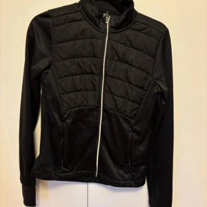 Svart quiltad träningsjacka med dragkedja - Säljer en svart träningsjacka med quiltade partier framtill och hel dragkedja. Jackan har hög krage, långa ärmar och två fickor med dragkedja. Materialet är slätt och lätt, perfekt för utomhusaktiviteter. Reflekterande prickar baktill för extra synlighet.