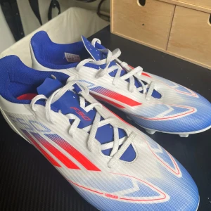 Adidas F50 fotbollsskor blå/vit/röd - Säljer ett par Adidas F50 fotbollsskor i en riktigt clean färgkombo av blått, vitt och rött. Skorna har snörning, platt sula med fasta dobbar och är tillverkade i syntetmaterial med texten 'F50' och 'Fiberskin' på sidan. Perfekta för dig som vill sticka ut på planen. Skorna har används en gång. Nypris är 600kr