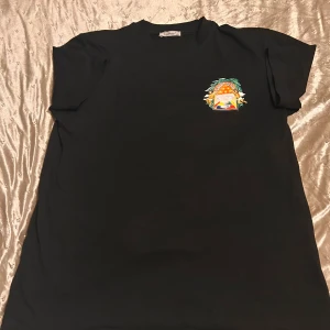 Svart t-shirt från Casablanca - Svart t-shirt från Casablanca med färgglatt tryck av änglar och fruktskål på ryggen och mindre tryck på bröstet. Klassisk passform och rund hals. Materialet känns mjukt och skönt, perfekt för en chill look. Storlek xxl men sitter som L