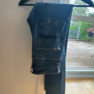 Blå jeans från Nudie Jeans, slim fit - Säljer ett par klassiska blå jeans från Nudie Jeans med snyggt slitna detaljer och kontrastsömmar. Modellen är Grim Tim som har smal passform, fem fickor och Nudie-loggan på bakfickan. Perfekta för dig som gillar en stilren och avslappnad look.