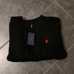 Svart stickad Polo Ralph Lauren tröja - Svart kabelstickad tröja från Polo Ralph Lauren med klassisk rund halsringning och röd broderad logga på bröstet. Tröjan har långa ärmar och ribbade muddar vid ärmslut och nederkant. Perfekt för en stilren och tidlös look.