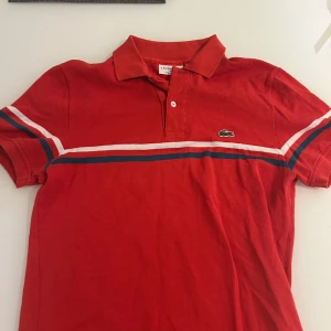 Röd Lacoste pikétröja med ränder - Röd pikétröja från Lacoste med klassisk krage och knäppning framtill. Tröjan har vita och mörkblå ränder över bröstet och ärmarna samt den ikoniska gröna krokodilloggan på bröstet. Tillverkad i bomull och har korta ärmar.