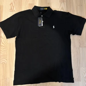 Svart Ralph Lauren Polo - Svart pikétröja från Polo Ralph Lauren med klassisk krage, korta ärmar och vit broderad logga på bröstet. Tröjan är i bomull och har knappar framtill. Perfekt för en clean och stilren look.