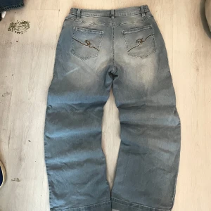 Ljusblå wide jeans från bpc - Säljer ett par ljusblå jeans från bpc med breda ben och broderade detaljer på bakfickorna. Jeansen har elastisk midja, klassisk knappstängning och dekorativa fickor fram. Perfekta för dig som gillar en avslappnad och trendig look.
