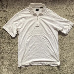 Vit t-shirt med krage från Jack & Jones - Vit t-shirt från Jack & Jones Premium i bomull, med klassisk krage och half zip-dragkedja framtill. Enkel och stilren design, perfekt för en clean look. Kortärmad modell som passar till många outfits.