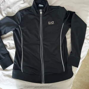 Svart EA7 ziptröja med vita detaljer - Svart ziptröja från EA7 Emporio Armani med vita ränder längs sidorna och dragkedja framtill. Hög krage, långärmad och logga både fram och bak. Snygg och sportig modell i mjukt syntetmaterial, perfekt till träning eller chill.