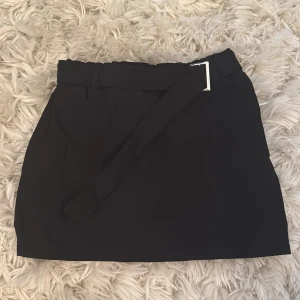 Svart kortkjol med bälte från Boohoo - Stilren svart kortkjol från Boohoo med brett tygbälte och silverfärgat spänne i midjan. Kjolen har en enkel, rak passform och är perfekt för dig som gillar minimalistisk stil. Tillverkad i ett mjukt syntetmaterial som känns bekvämt hela dagen. Aldrig använd