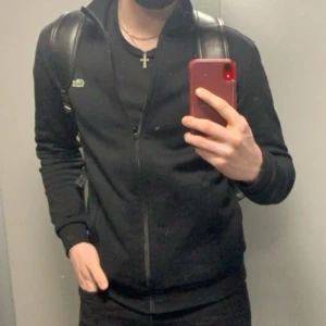 Lacoste Sport Men's SH7616 tröja - Svart långärmad ziptröja från Lacoste med klassisk krokodillogga på bröstet. Tröjan har hög krage, dragkedja hela vägen och ribbade muddar. Tillverkad i mjuk bomullsmix som är skön att bära. Perfekt för en clean och sportig look.