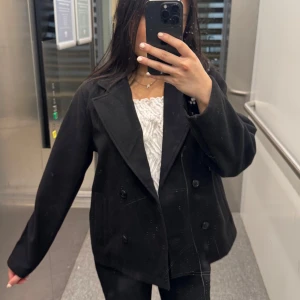 Svart kavaj med knappar - Stilren svart kappa med dubbelknäppning och bred krage. Jackan har en rak passform och är tillverkad i ett mjukt material som känns lite ulligt. 