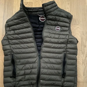 Olivgrön dunväst från Colmar - Snygg mörkgrön dunväst från Colmar Originals med quiltad design och två praktiska fickor med dragkedja. Västen har en hög krage och Colmar-logga på bröstet. Perfekt att slänga över en hoodie för extra värme när det är kyligt ute.