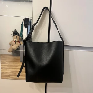 Svart axelväska med justerbart band - Superfin och elegant väska från Lindex.  Läderimitation med stort fack och två mindre fack inuti.  Använd max 5 gånger så den är i superfint skick! Säljes då det inte finns plats i garderoben och den inte kommer till användning! Höjd: 34cm. Längd: 31cm. Bredd: 13cm.