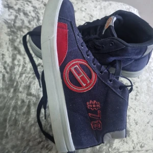 Mörkblå sneakers med röd detalj - Snygga mörkblå sneakers med röda broderade detaljer och logga på sidan samt #78. Skorna har snörning, vit sula och är tillverkade i ett mockaliknande material. Perfekta för dig som vill sticka ut med en sportig och unik stil.