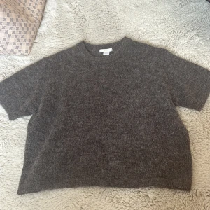 Brun stickad tröja från Gina Tricot - Mysig brun stickad tröja från Gina Tricot med kort ärm och rund hals. Tröjan har en lite boxig passform och är gjord i ett fluffigt, mjukt material som känns perfekt för kyligare dagar. Enkel och stilren design som funkar till många outfits.