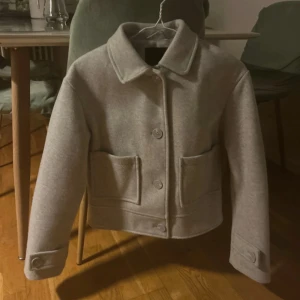 Grå ulljacka med stora fickor - Snygg grå jacka i ull med klassisk krage och stora framfickor. Jackan har knäppning med stora knappar framtill och på ärmsluten. Rak och boxig passform som ger en modern look.