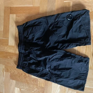 Svarta shorts från C.P. Company - Svarta shorts från C.P. Company med elastisk midja och snörning. Tillverkade i ett lätt syntetmaterial, har dragkedjeförsedda fickor och den klassiska C.P. Company-linsen på benet. Perfekta för en chill och sportig stil.