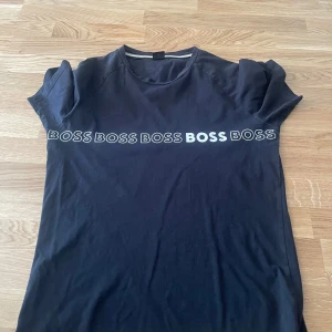 Svart BOSS t-shirt i bomull - Svart t-shirt från BOSS i ekologisk bomull med slim fit-passform. Framsidan har flera BOSS-loggor tryckta i vitt över bröstet. Klassisk rund halsringning och korta ärmar. Perfekt för en clean och stilren look.