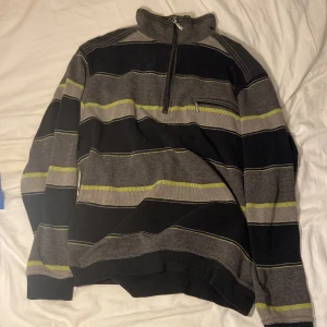 Randig halvzip tröja i grått och svart - Randig tröja med halv dragkedja i halsen och liten ficka med dragkedja på bröstet. Färgerna är svart, grå och inslag av limegrönt. Tröjan har lång ärm och är stickad i ett mjukt material. Perfekt för kyligare dagar och har en chill vibe.