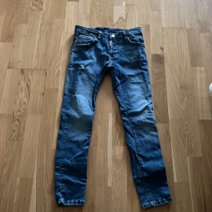 Blå jeans från Dsquared2, storlek 14Y - Snygga blå jeans från Dsquared2 med slitna detaljer och klassisk femficksdesign. Jeansen har en rak passform och är tillverkade i en mix av bomull, polyester och elastan för skön stretch. Perfekta för dig som gillar en cool och avslappnad stil.