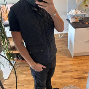 Svart quiltad väst från Belstaff - Snygg svart quiltad väst från Belstaff med hög krage och två stora fickor framtill. Perfekt för lager-på-lager. Nypris ligger på en bit över 2000kr, mitt pris är 699! Perfekt nu till hösten samt våren nästa år! Västan är i storlek M/L.