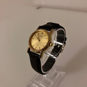 Citizen Quartz klocka med svart läderband - Stilren analog klocka från Citizen med guldfärgat runt ur och svart läderband. Urtavlan är enkel och guldig med tunna svarta markeringar och diskret datumfönster. Klassisk design som passar dig som gillar tidlös stil. Behöver ett nytt batteri.