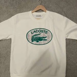  Lacoste tröja - Vit sweatshirt från Lacoste med stor grön krokodil-logga och text på bröstet. Klassisk rund halsringning och ribbade muddar. Perfekt för dig som gillar stilren design med sportig touch.