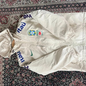 Vit Nike Brasil vindjacka med huva - Vit vindjacka från Nike med Brasil-logga och sponsormärken på bröstet och ärmarna. Jackan har huva, dragkedja framtill och elastiska muddar. Perfekt för dig som gillar fotboll och vill ha en sportig stil.