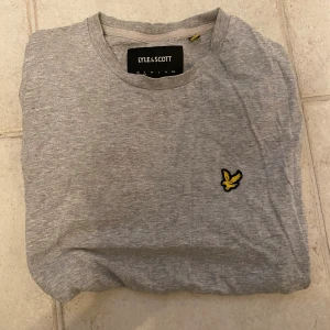 Grå t-shirt från Lyle & Scott - Snygg grå t-shirt från Lyle & Scott med klassisk rund hals och korta ärmar. På bröstet sitter den ikoniska gula fågeln broderad. T-shirten är gjord i mjuk bomull och har en clean, enkel look som funkar till allt.
