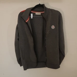 Grå ziptröja från Moncler S - Mörkgrå ziptröja från Moncler med broderad logga på bröstet och röda/vita detaljer i kragen. Tröjan har hög krage, långa ärmar och två fickor framtill. Perfekt för dig som gillar stilrena och sportiga plagg. Den är lite smutsig på fram sidan. Men det går bort i tvötten