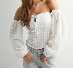 Vit offshoulder blus - Supersöt vit blus med offshoulder och långa puffärmar. Toppen har ett diskret strukturmönster, knytning framtill och smockad resår vid ärmslut och axlar. Perfekt för en trendig och romantisk look. Säljer då den aldrig kommit till användning! Säljs inte längre