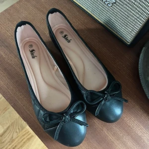 Svarta ballerinaskor med rosett från SheIn - Stilrena svarta ballerinaskor från SheIn med rund tå och söt rosettdetalj framtill. Skorna har en platt sula med blommönster undertill och är tillverkade i syntetmaterial. Perfekta för dig som gillar en klassisk och enkel look.