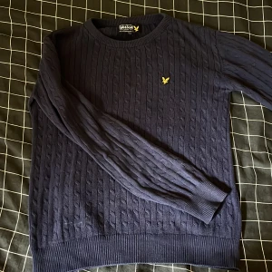 Mörkblå kabelstickad tröja Lyle & Scott - Kabelstickad mörkblå tröja från Lyle & Scott med rund hals och klassisk gul logga broderad på bröstet. Tröjan har långa ärmar och ribbade muddar vid ärmslut och nederkant. Perfekt för dig som gillar stilren och tidlös design.
