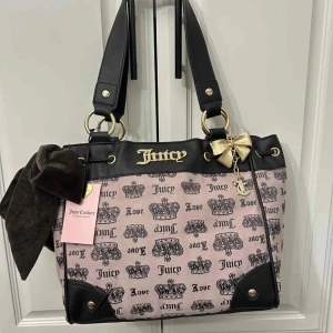Juicy Couture daydreamer  handväska med rosett - Superfin handväska från Juicy Couture i rosa och svart med kronmönster, guldfärgade detaljer och en stor brun sammetsrosett. Väskan har axelremmar, innerfack med dragkedja och snyggt Juicy Couture-tryck även på insidan. Perfekt för dig som gillar statement-accessoarer.