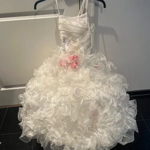 Vit prinsessklänning med volanger - Superfin långklänning för barn  i vitt med massor av volanger och glansigt tyg. Klänningen har smala axelband, dekorativa blommor och pärlor upptill samt rosa blommor i midjan. Perfekt blomsterflicka/ brudnäbb för bröllop