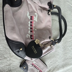 Juicy Couture rosa axelväska & plånbok - Säljer en superfin rosa axelväska i mocka från Juicy Couture med svarta skinn-detaljer och guldfärgade metalldelar. Väskan har broderad logga i vitt och rosa samt ett hjärtformat svart hänge. Matchande plånbok medföljer, båda med dragkedja och coolt tryckt innerfoder.
