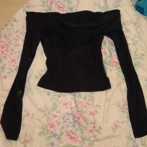 Svart ribbad offshoulder topp - Säljer en svart ribbad offshoulder topp med långa ärmar. Snygg passform som sitter tight och markerar axlarna. Perfekt för dig som vill ha en stilren och trendig look. Materialet är stretchigt och bekvämt.