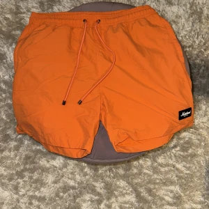 Orange badshorts från SQRTN Company - Säljer ett par orange badshorts från SQRTN Company i storlek M. Shortsen har elastisk midja med snörning, meshfoder och en liten svart logotyp på framsidan. Perfekta för sommaren och strandhäng, riktigt skön passform och stilren design.