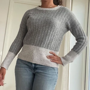 Grå och vit stickad tröja - Säljer en ribbstickad tröja i grått och vitt med rund halsringning. Tröjan har långärmade ärmar och en bredare vit kant vid nederdelen och ärmsluten. Perfekt för dig som gillar en enkel men snygg look.