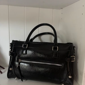 Svart handväska - Snygg svart handväska i glansigt skinn med silverfärgade metalldetaljer och dragkedja framtill. Väskan har två stadiga handtag och coola spänndetaljer på sidorna. Perfekt för dig som vill ha en stilren accessoar till din outfit. Det finns ett oanvänt långt band.