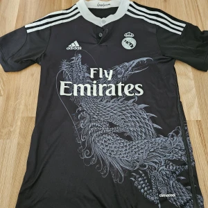 Real Madrid svart fotbollströja Adidas S - Svart Real Madrid fotbollströja från Adidas med vit krage och axelränder. Stort drakmotiv på framsidan, klubbmärke och Adidas-logga på bröstet samt Fly Emirates-tryck. Tillverkad i lätt och ventilerande climacool-material.